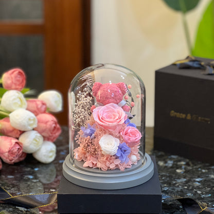 Pink teddy bear nestled in preserved roses glowing under a glass dome
