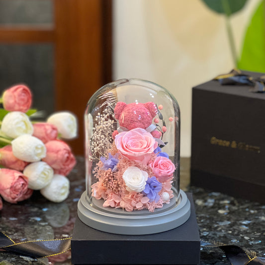 Pink teddy bear nestled in preserved roses glowing under a glass dome