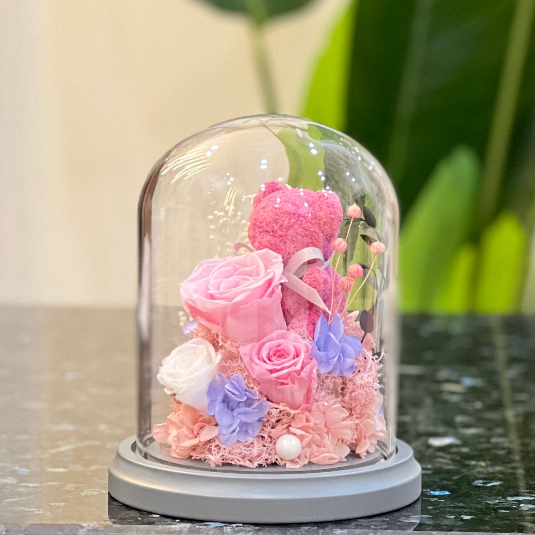 Side view of a pink rose bear with preserved roses and hydrangea in a glass dome
